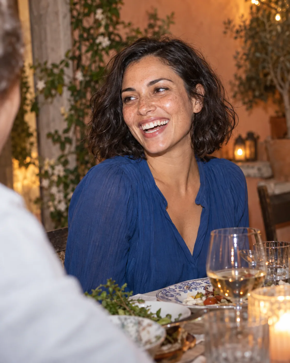 A woman in a blue blouse laughing with a friend at an outdoor dinner table