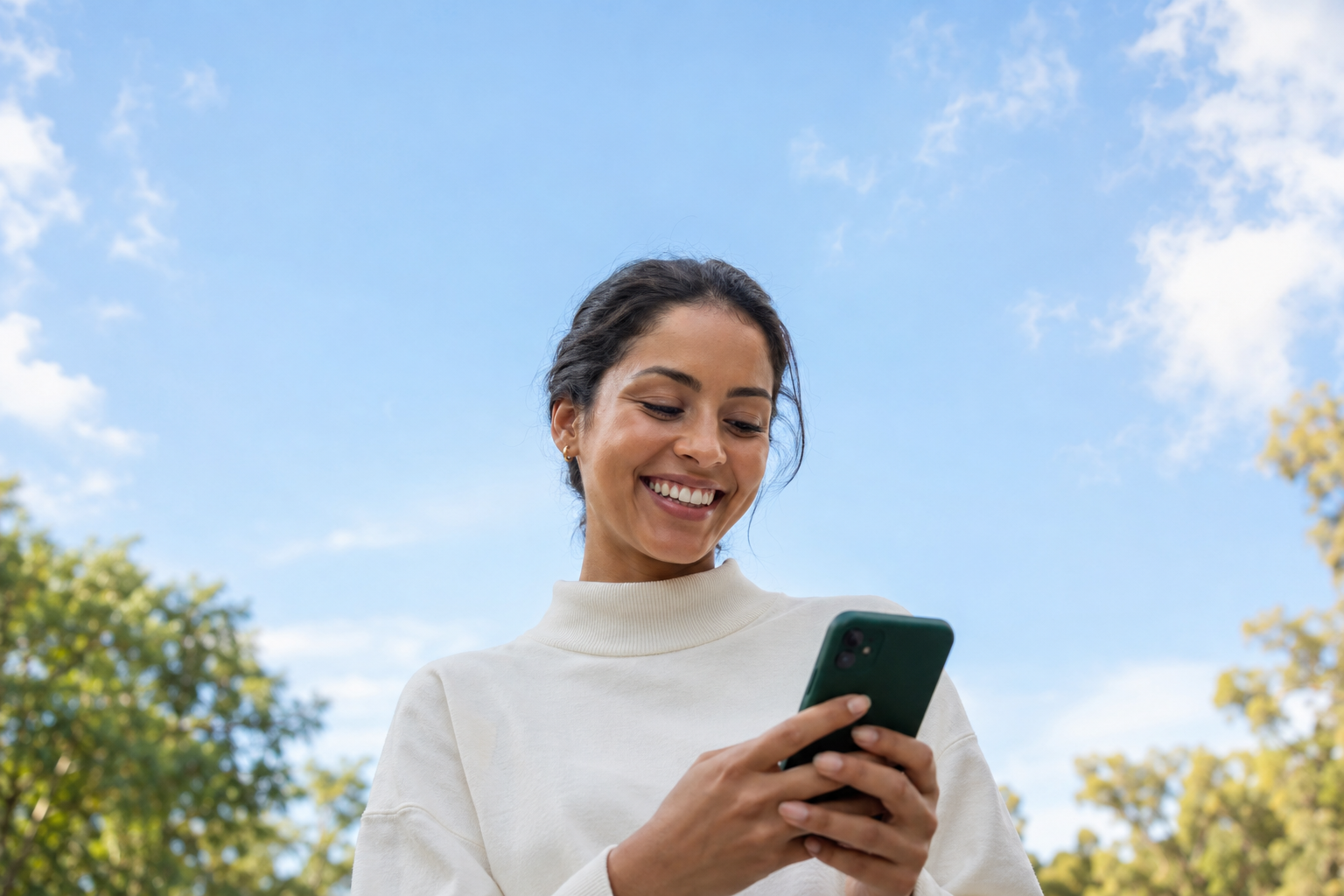A smiling woman in an ivory sweater looking at a dark green smartphone outdoors beneath a blue sky